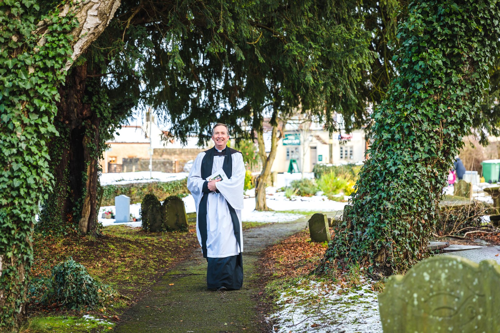 The vicar waits beneath an ivy-clad archway in the snowy churchyard
