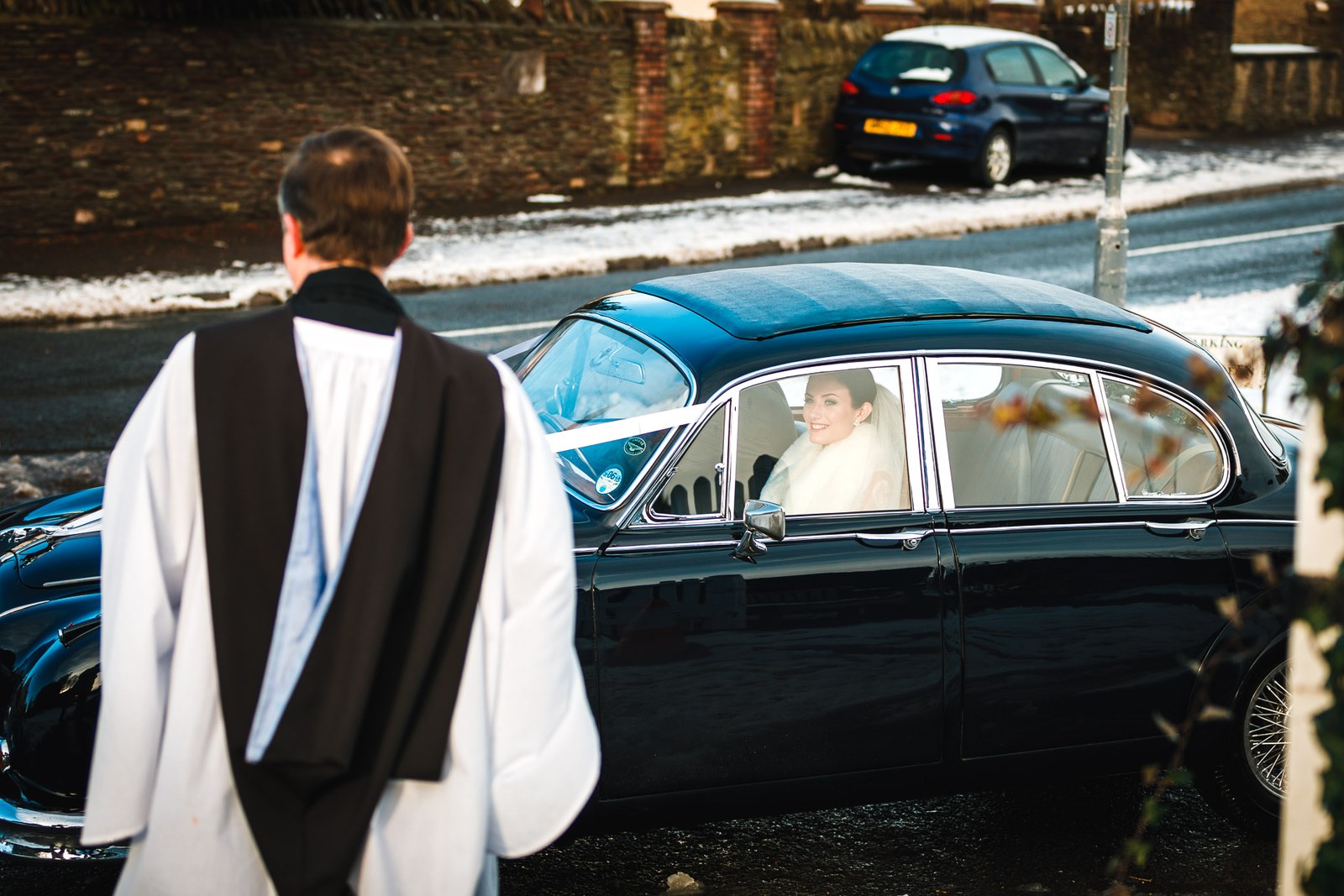 The vicar walks toward the ribboned vintage car carrying the bride through the snow