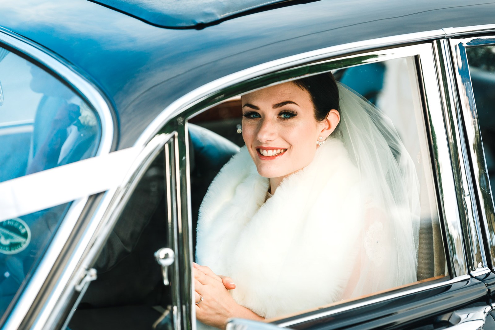 Bride in a white fur stole smiles from the window of the classic wedding car