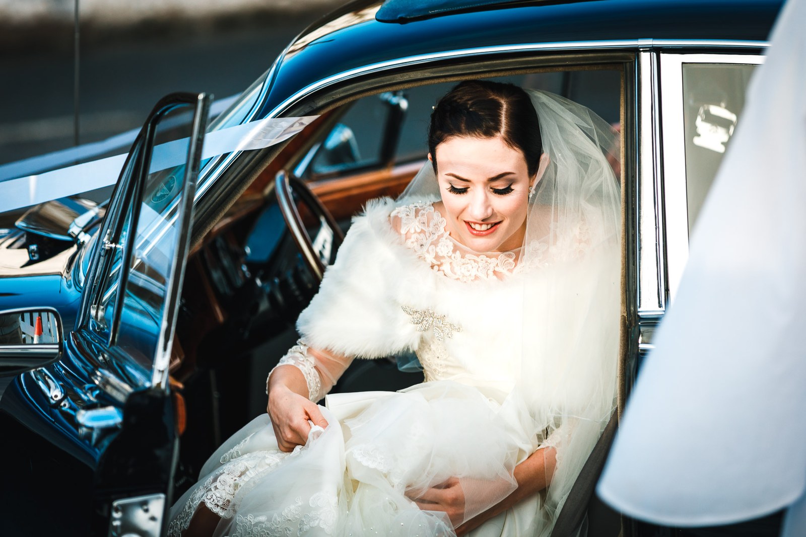 Bride steps from the vintage car, gathering her gown in her fur wrap
