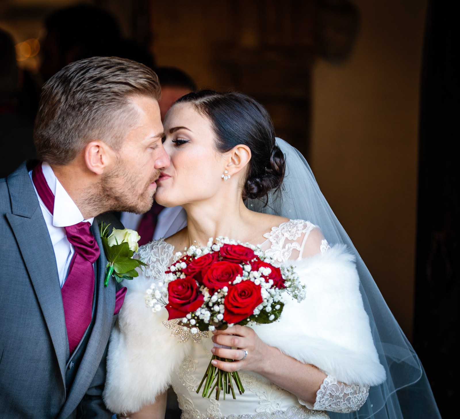 The groom kisses his bride, her red rose bouquet held between them