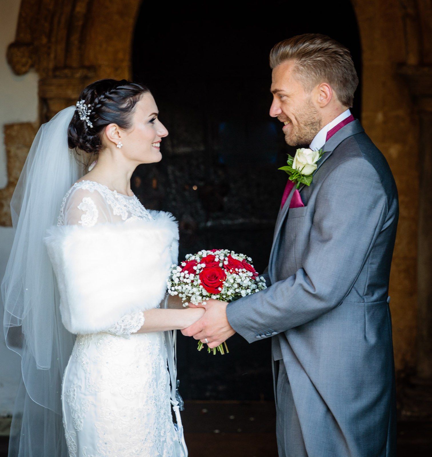 Bride and groom stand face to face, red roses cradled at her chest