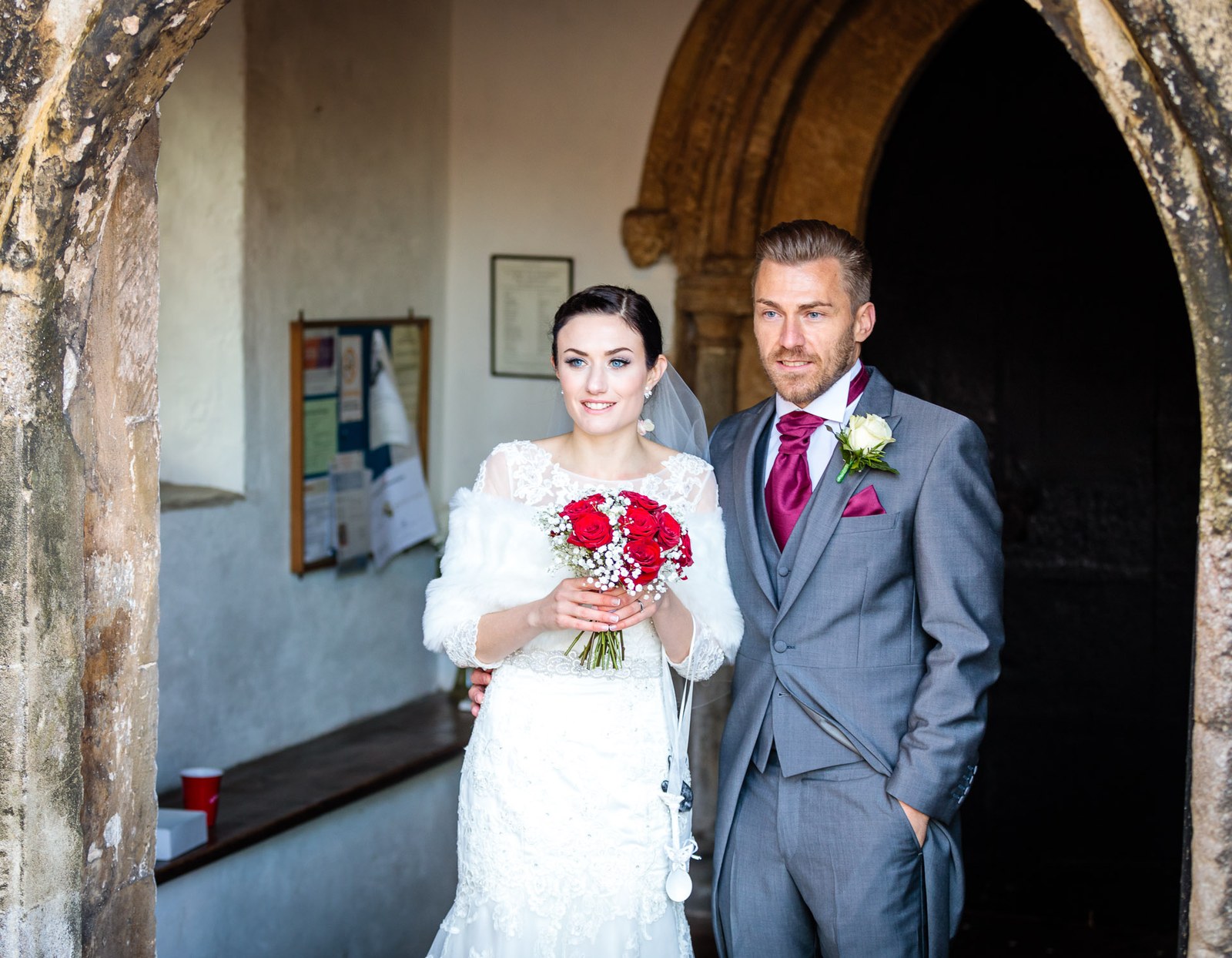 Bride holds her bouquet beside the groom in the stone church porch