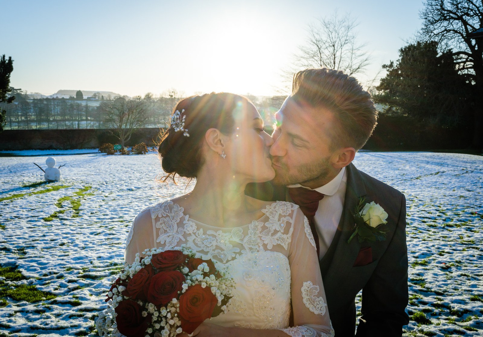 The couple kiss in the sunlit snow, a small snowman behind them