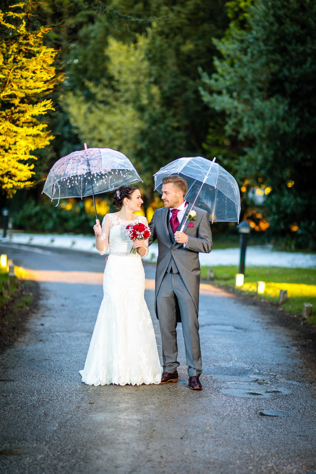 Bride and groom walk a lamp-lit drive sheltering under clear umbrellas