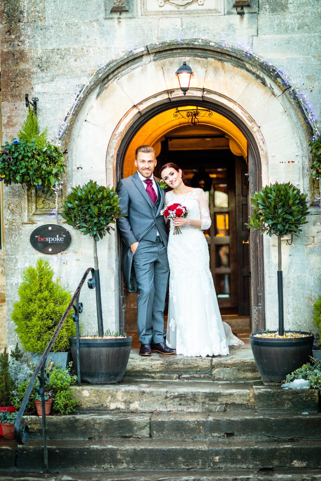 The couple stand between bay topiary in the arched Stonehouse Court Hotel entrance