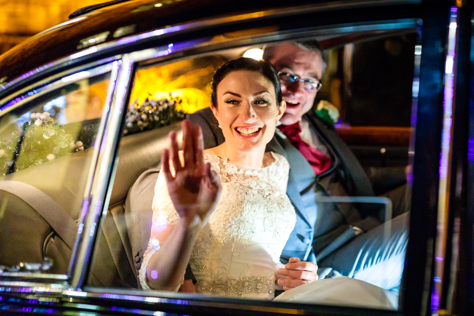 Bride waves from the vintage car window in the evening light