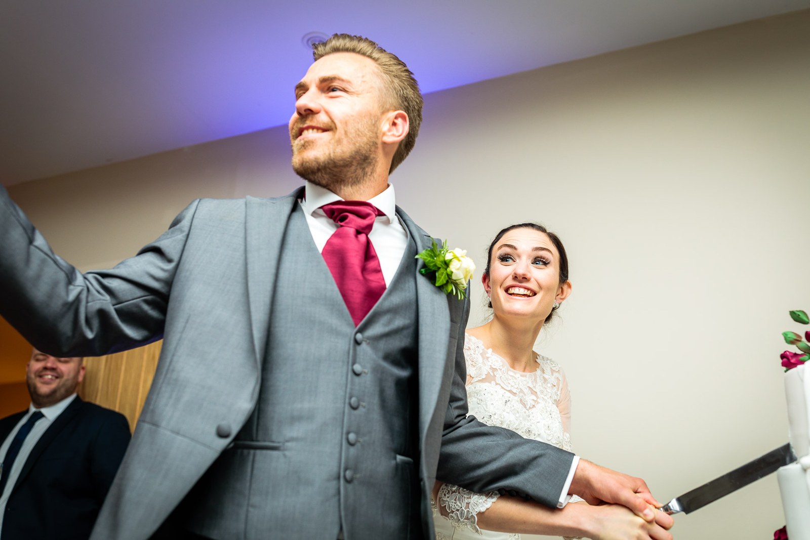 The groom flings an arm wide as the couple set the knife to the cake
