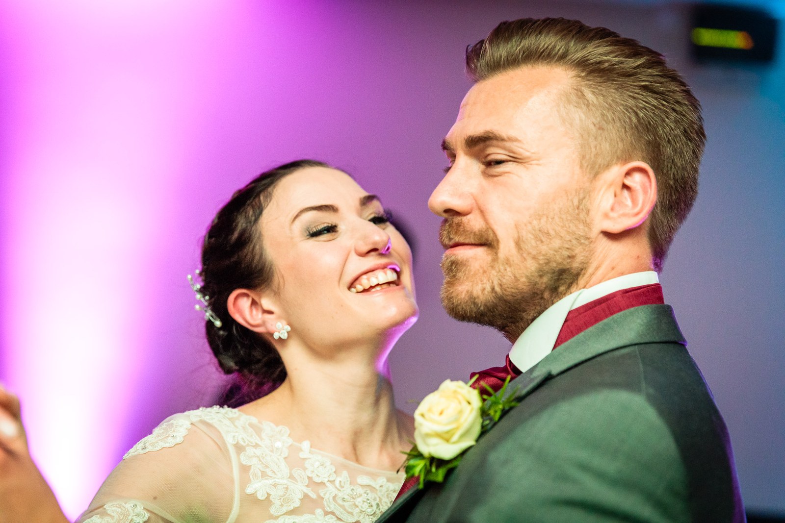 The bride laughs against the groom in their first dance beneath pink light
