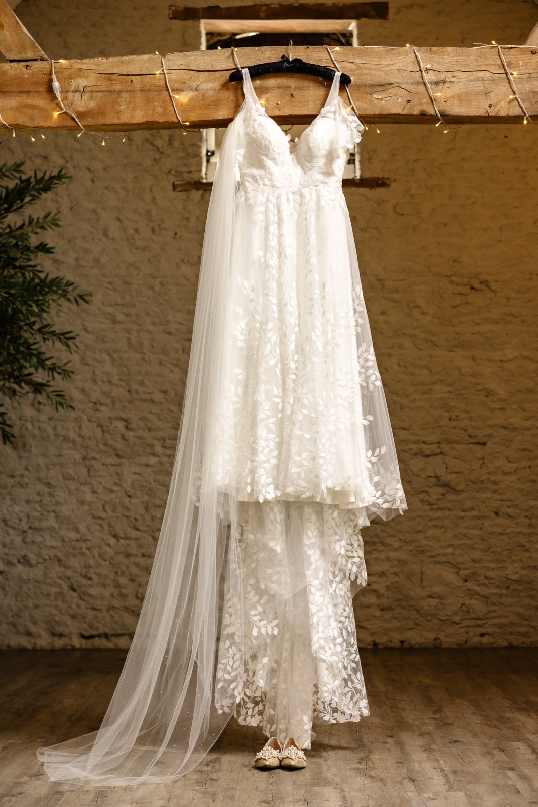 Lace wedding dress hanging from a timber beam against the stone wall at Stratton Court Barn.