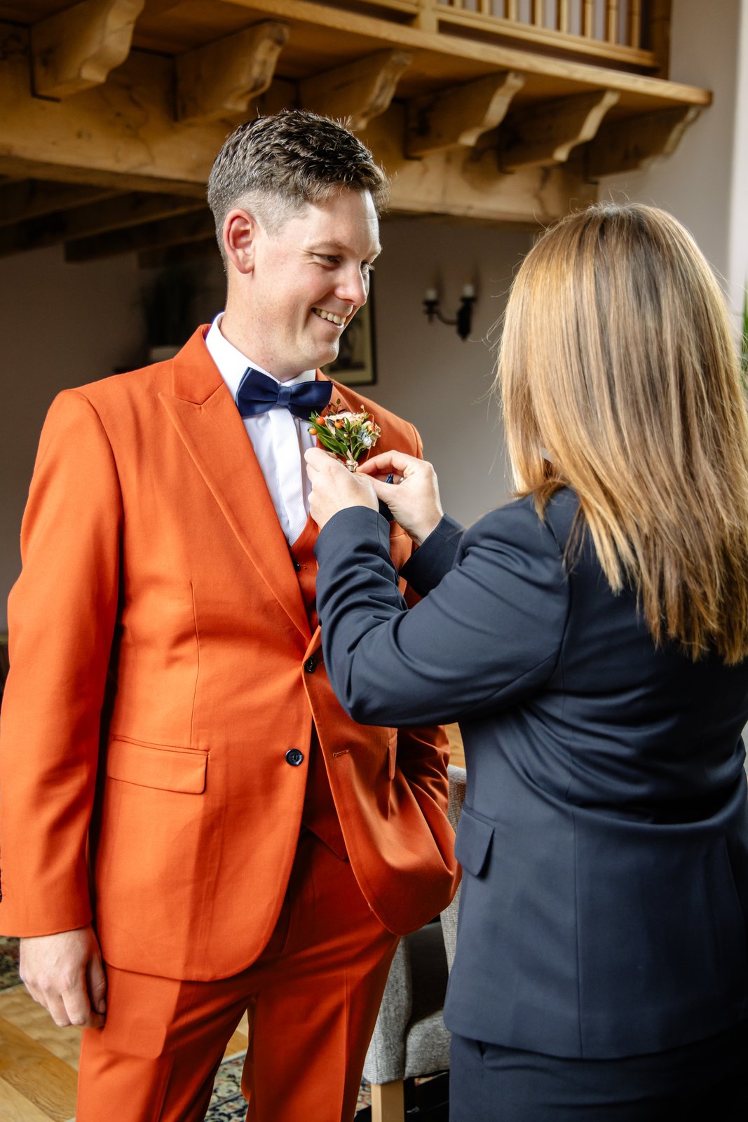 A woman pinning a buttonhole to the groom's burnt orange jacket in a beamed room.