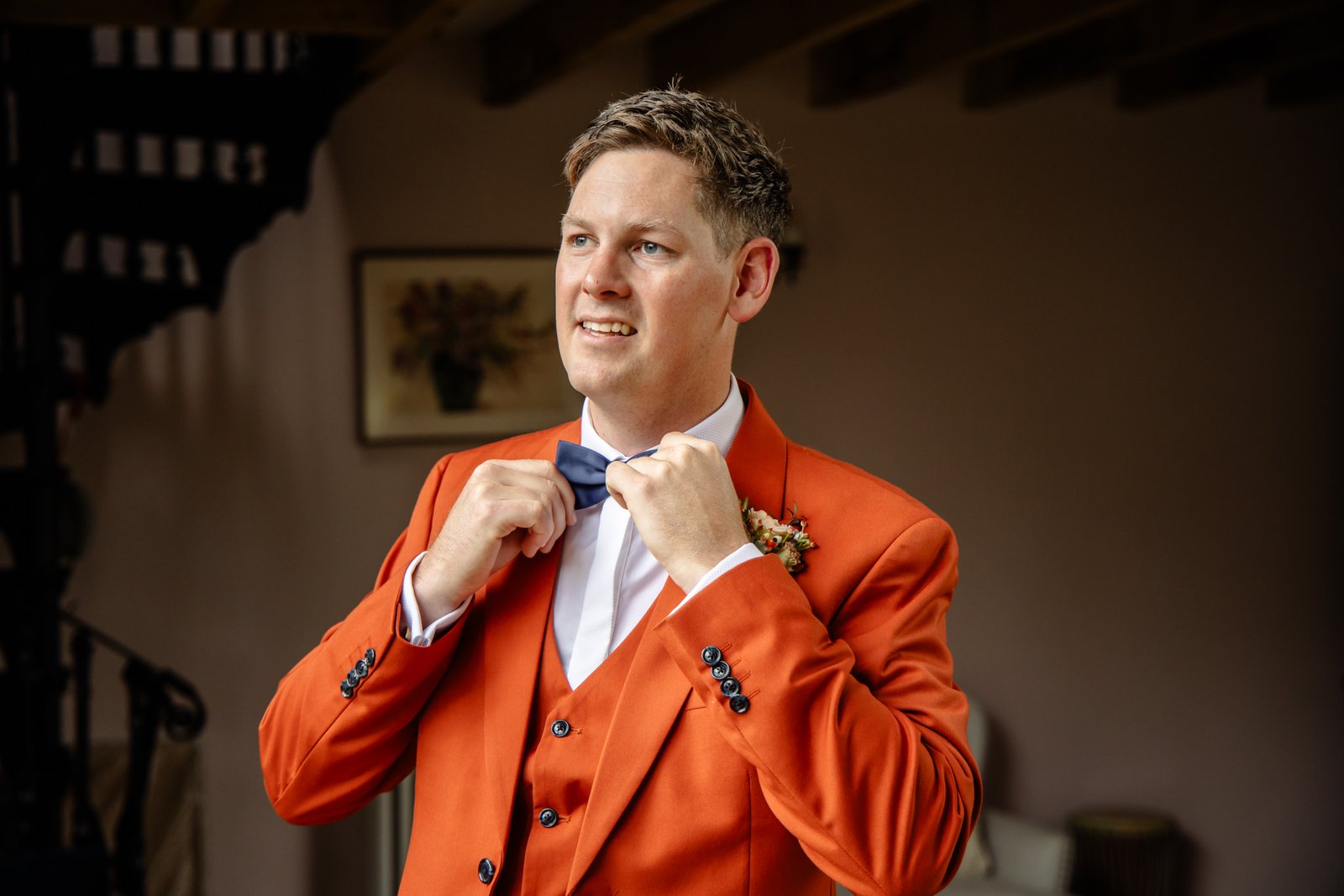 The groom straightening his navy bow tie, a dark wooden staircase behind him.