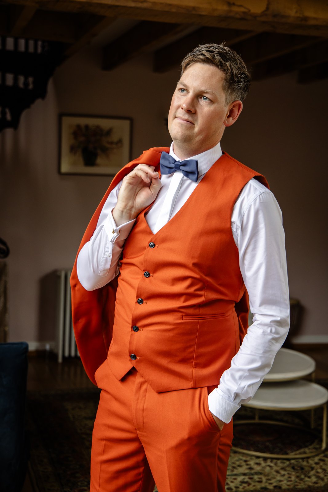 The groom in a burnt orange three piece suit fixing his cuff beside a window.