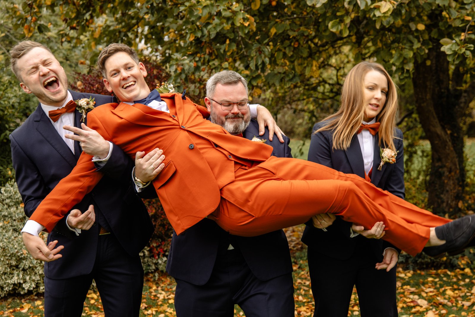 Groomsmen lifting the groom horizontally off the ground, everyone laughing, autumn trees behind.