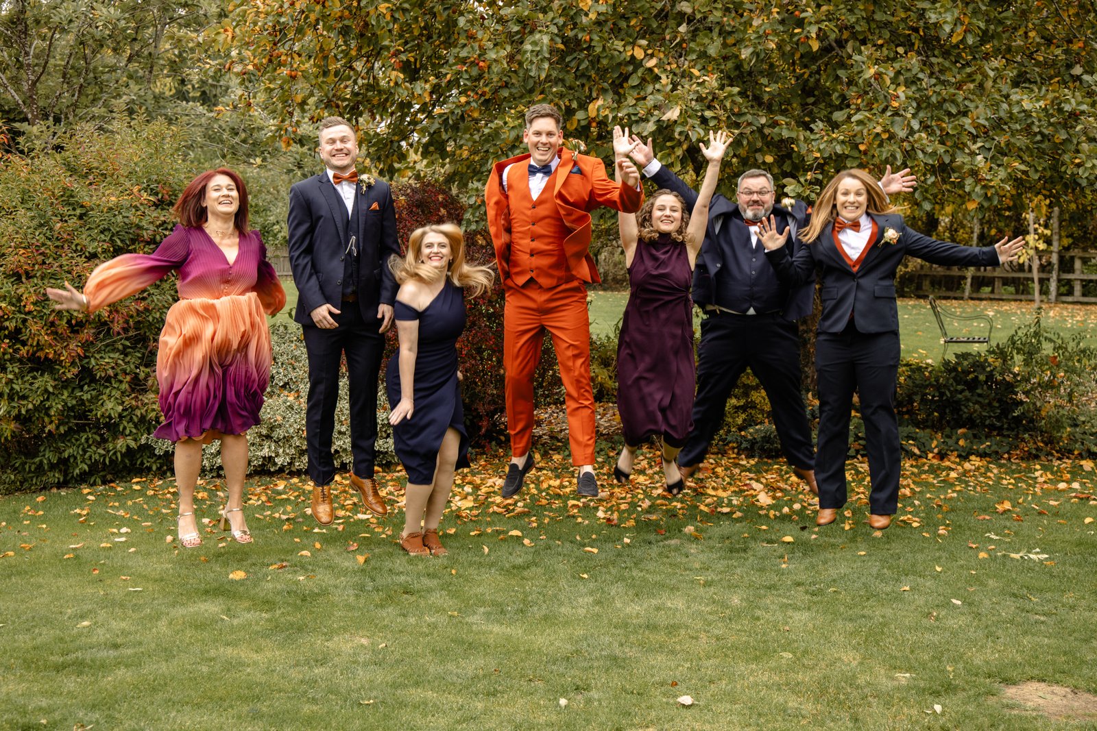 The wedding party jumping in the air on a lawn scattered with autumn leaves.