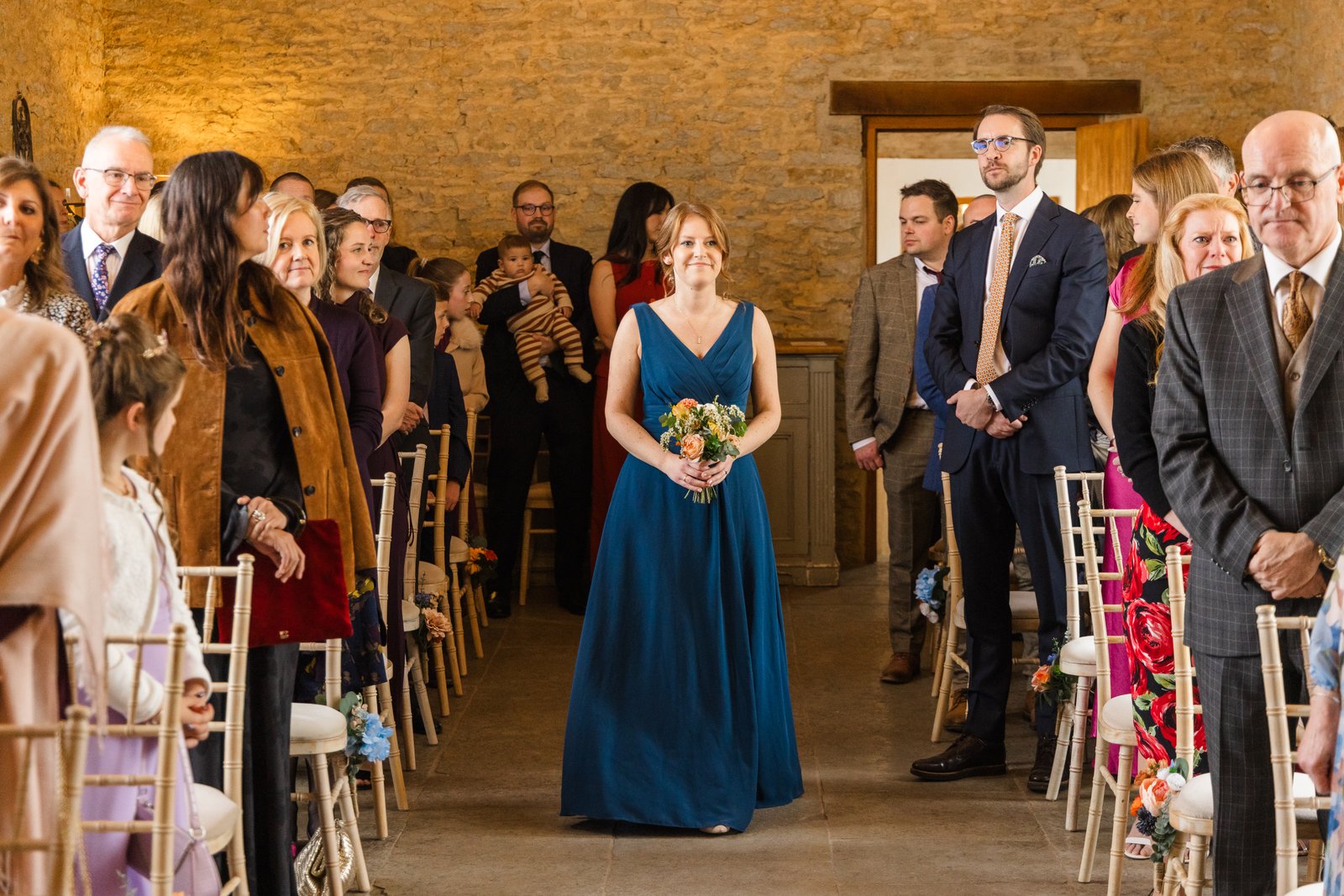 A bridesmaid in navy coming down the aisle between rows of guests at Stratton Court Barn.