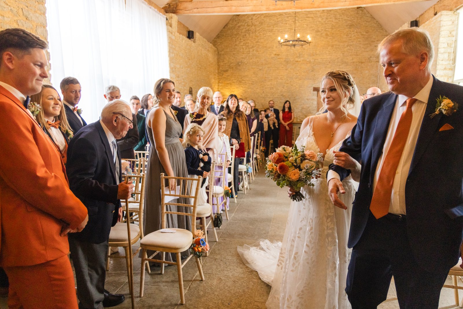The bride and her father walking between the guests, her peach and orange bouquet in hand.