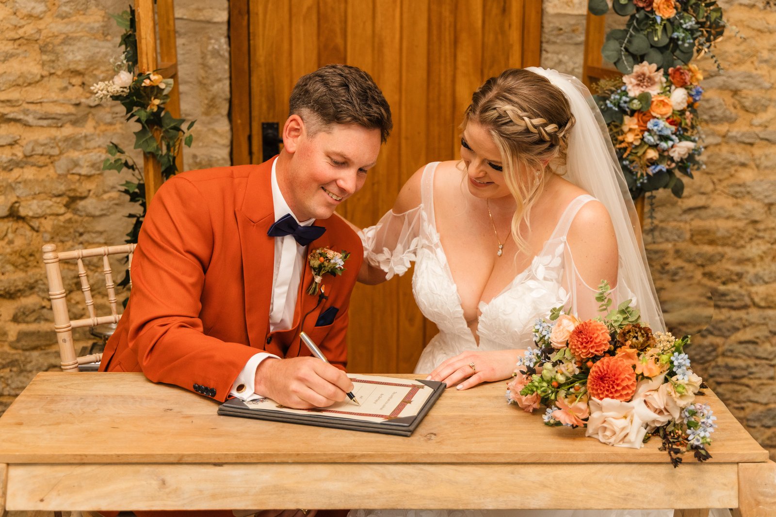 The couple signing the register at a wooden table, a floral wreath on the door behind them.