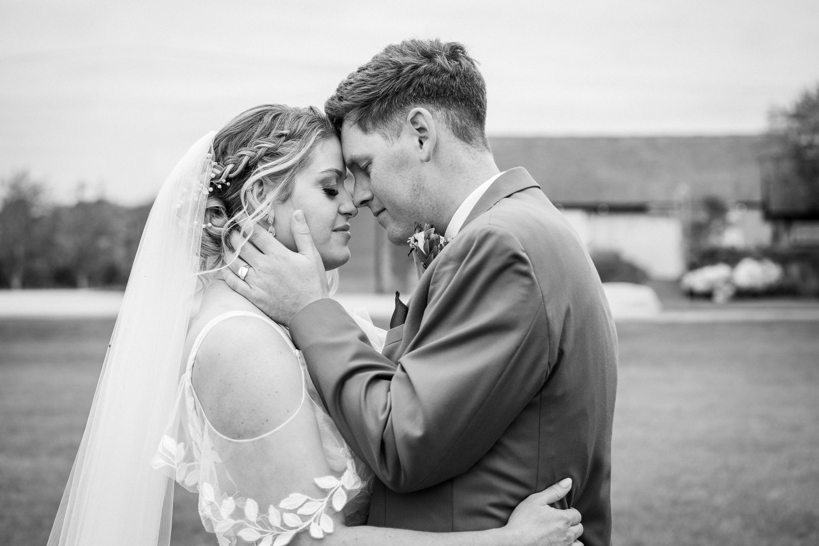 Black and white portrait of the couple embracing, the barn buildings in the distance.
