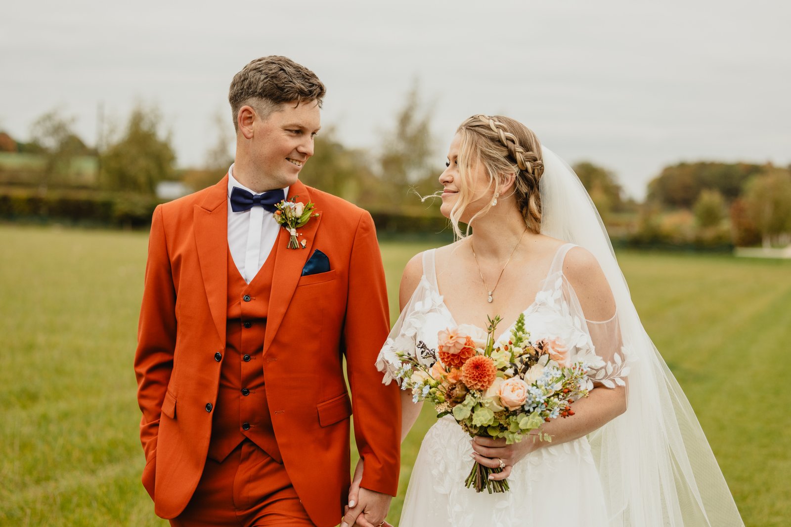 The bride and groom walking through the field beside Stratton Court Barn with her bouquet.