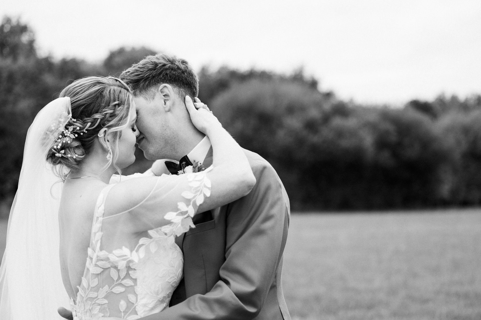 The bride and groom about to kiss in black and white, hedgerow and open grass behind.