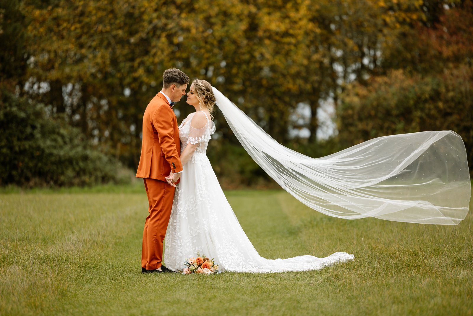 The bride's veil blown out sideways as the couple stand together under autumn trees.