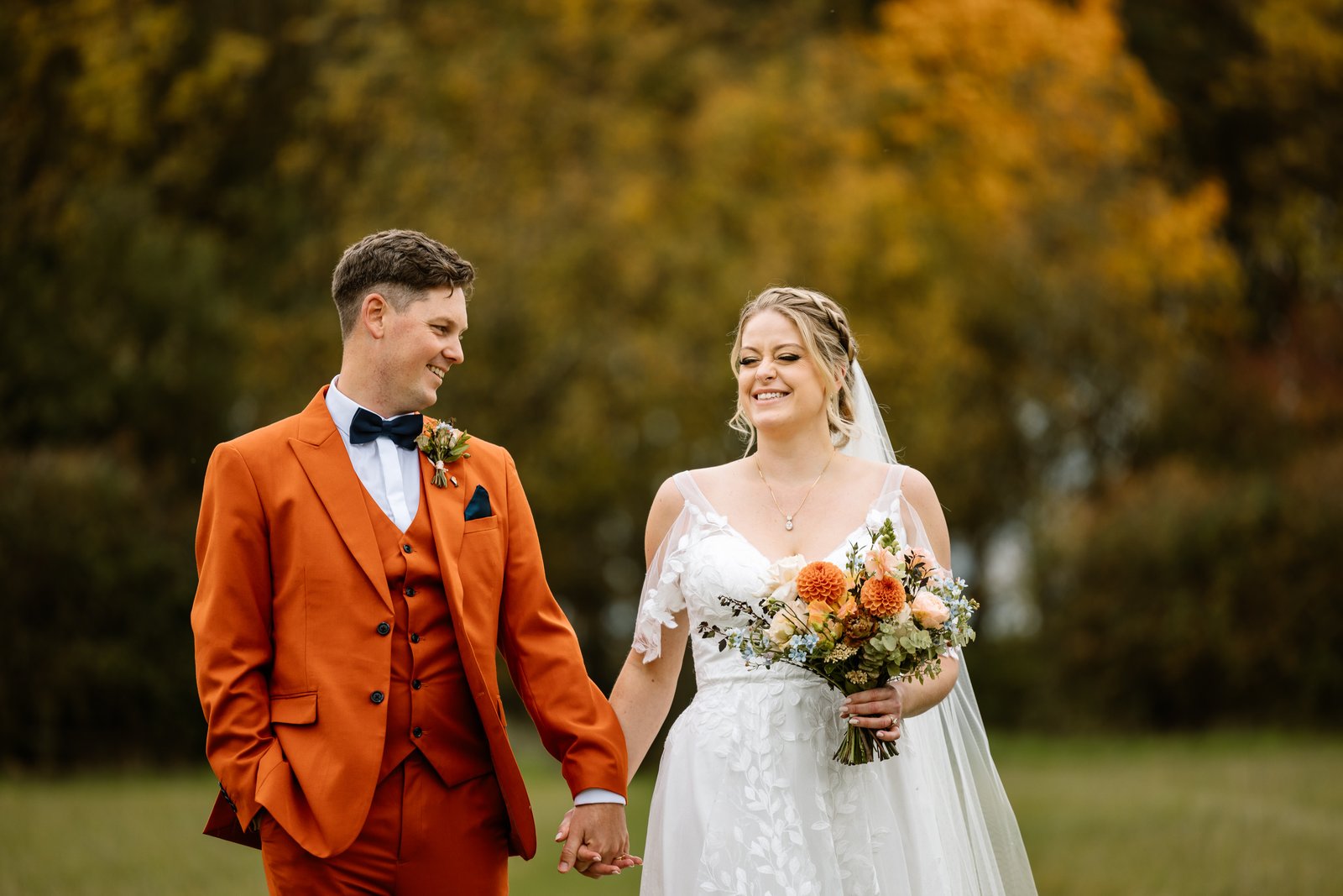 The couple walking hand in hand past golden autumn trees near Stratton Court Barn.