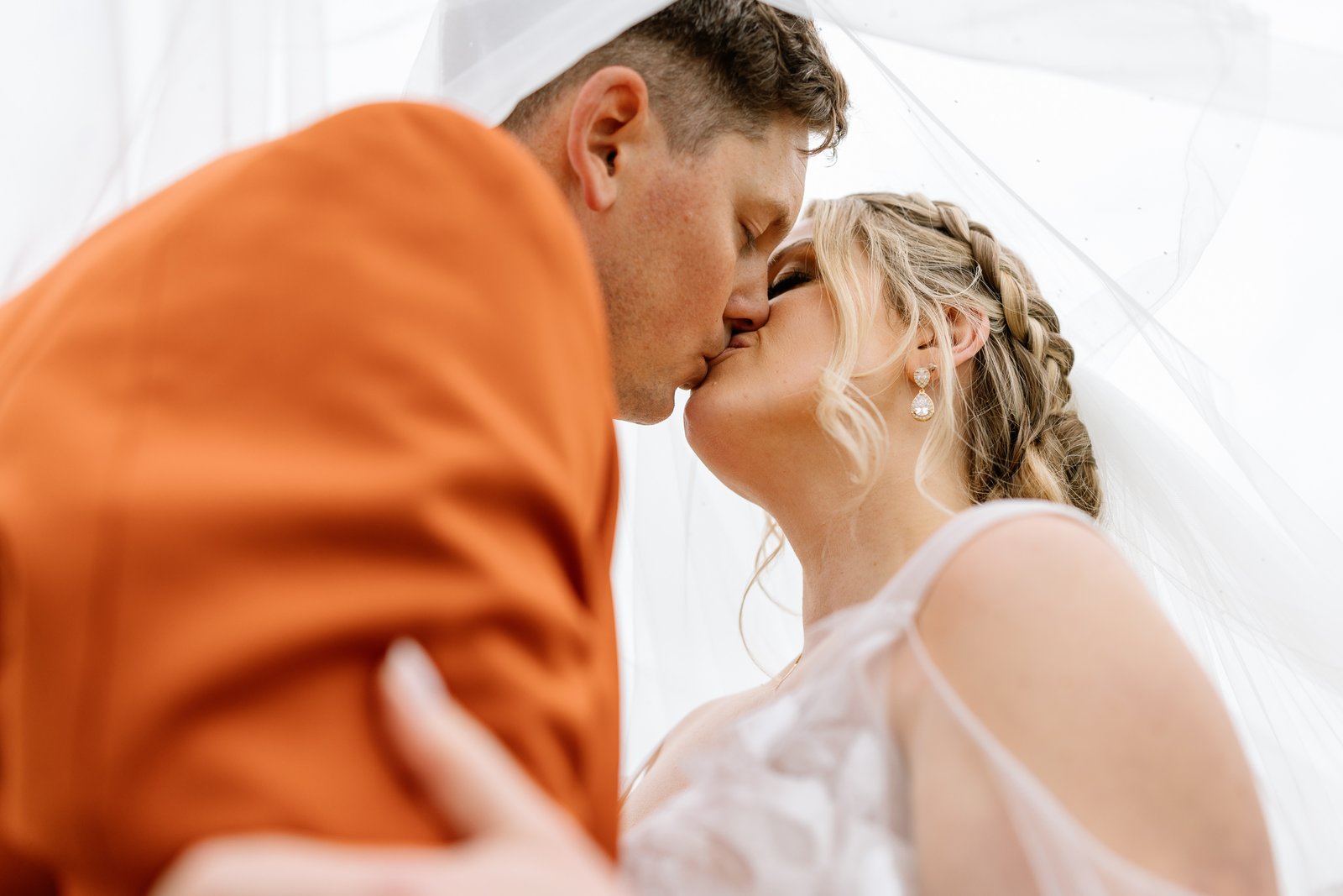 The bride and groom kissing beneath her veil, his orange sleeve around her shoulder.