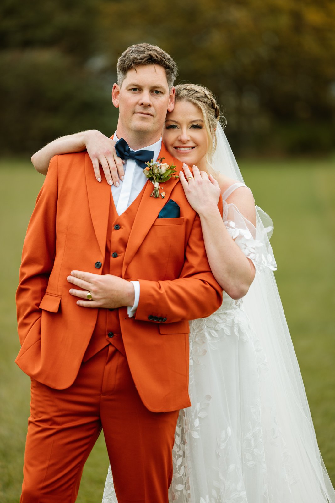 The bride with her arms around the groom's shoulders, autumn trees behind them.