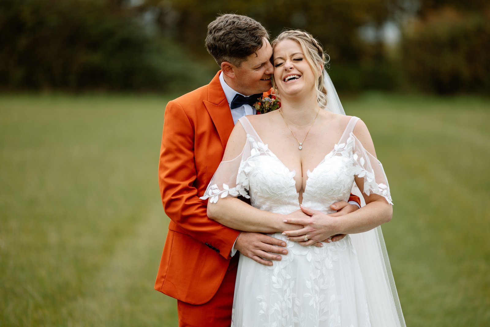 The groom holding the bride from behind as she laughs, open grass around them.