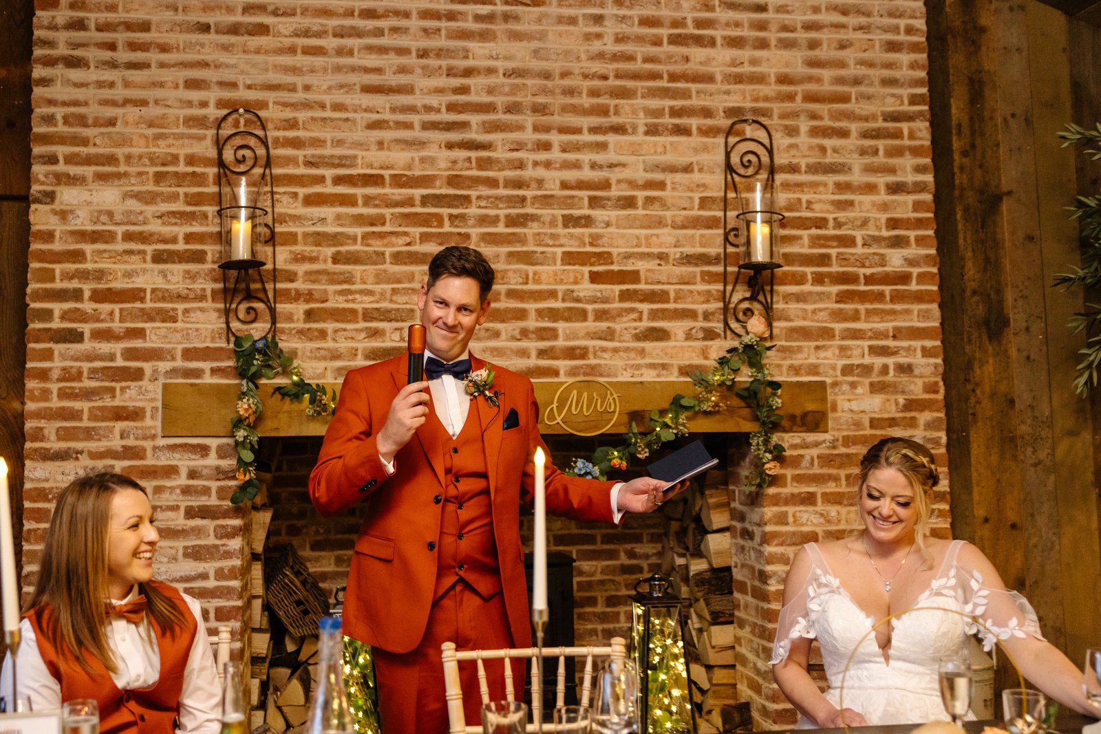The groom giving his speech beside the fireplace as the bride listens from the table.