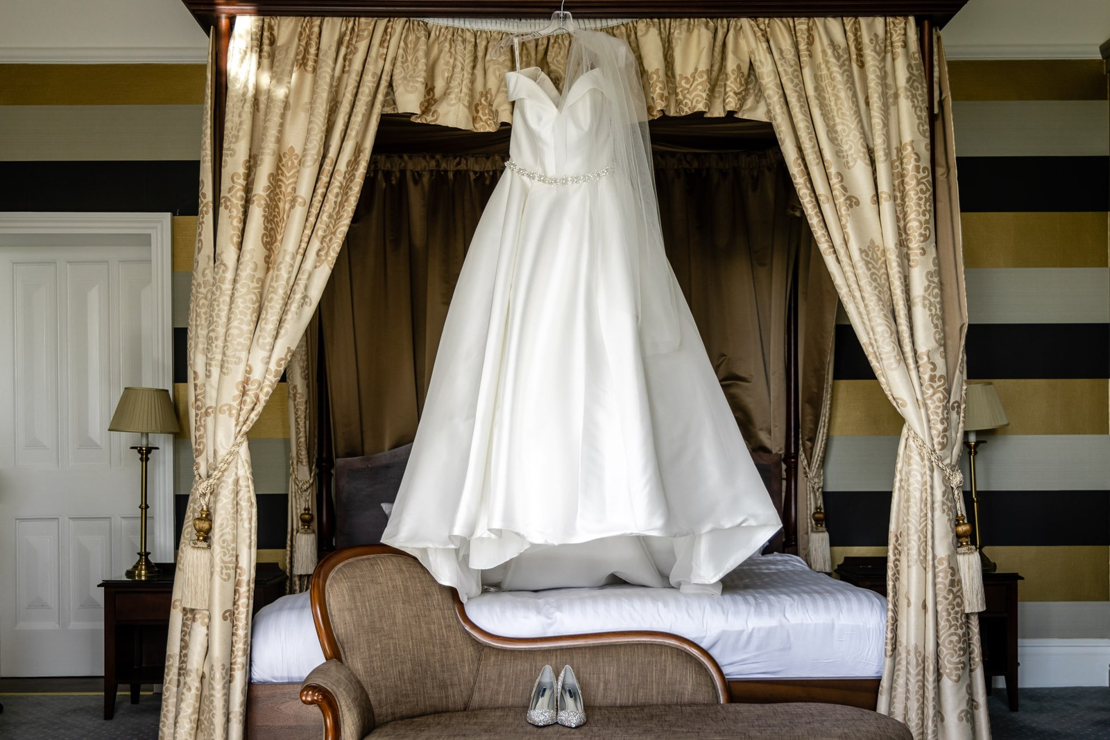 The wedding gown hanging from a four-poster bed with bridal shoes on the chaise below