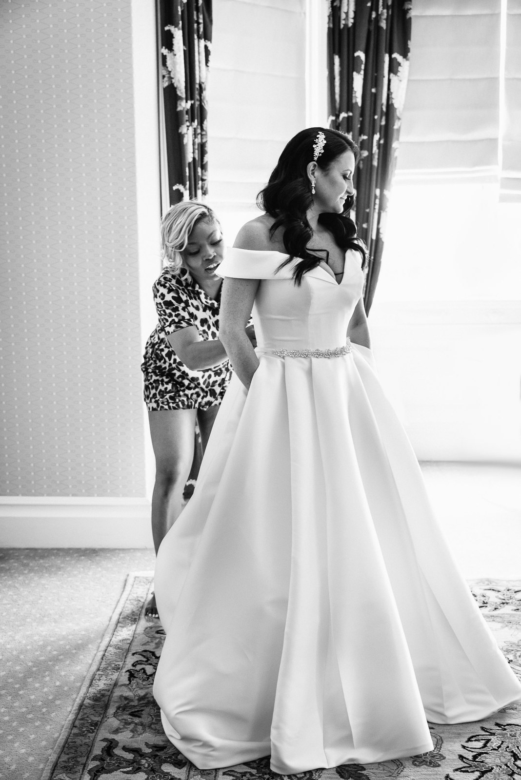 Black-and-white shot of a helper doing up the bride's dress by a window