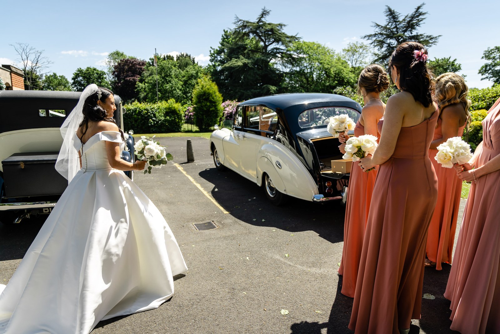 Bride greeting her bridesmaids in rust dresses beside a vintage wedding car