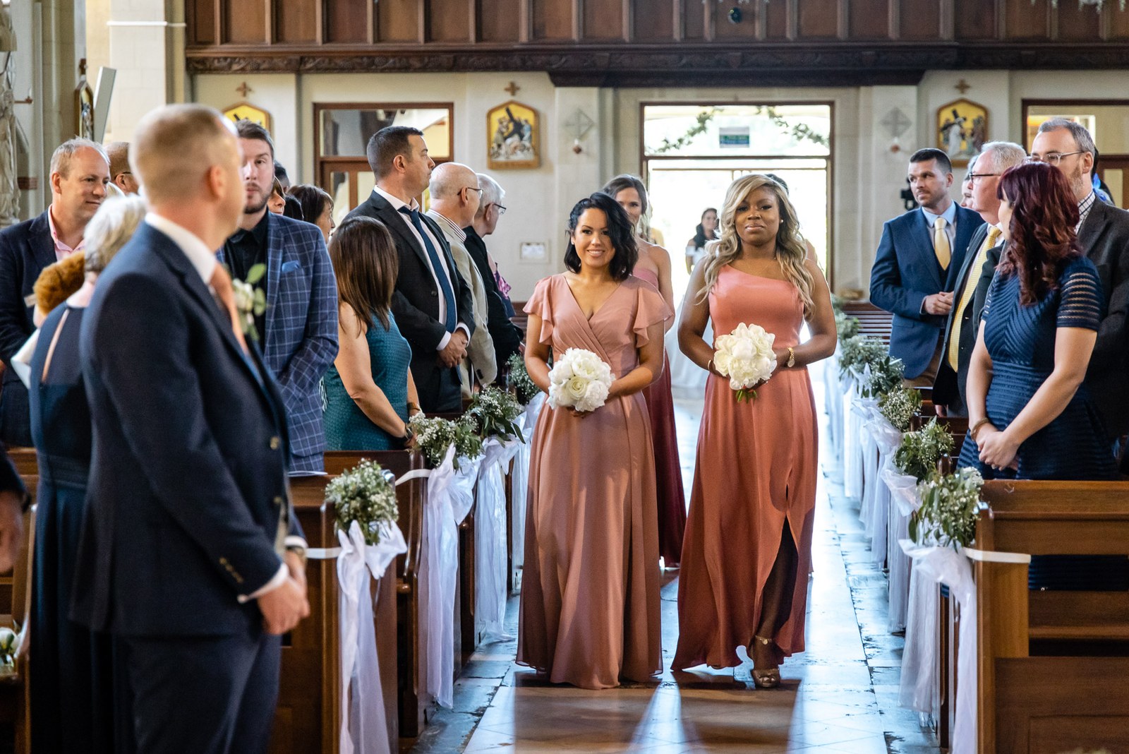 Bridesmaids in rust gowns walking down the church aisle with white bouquets