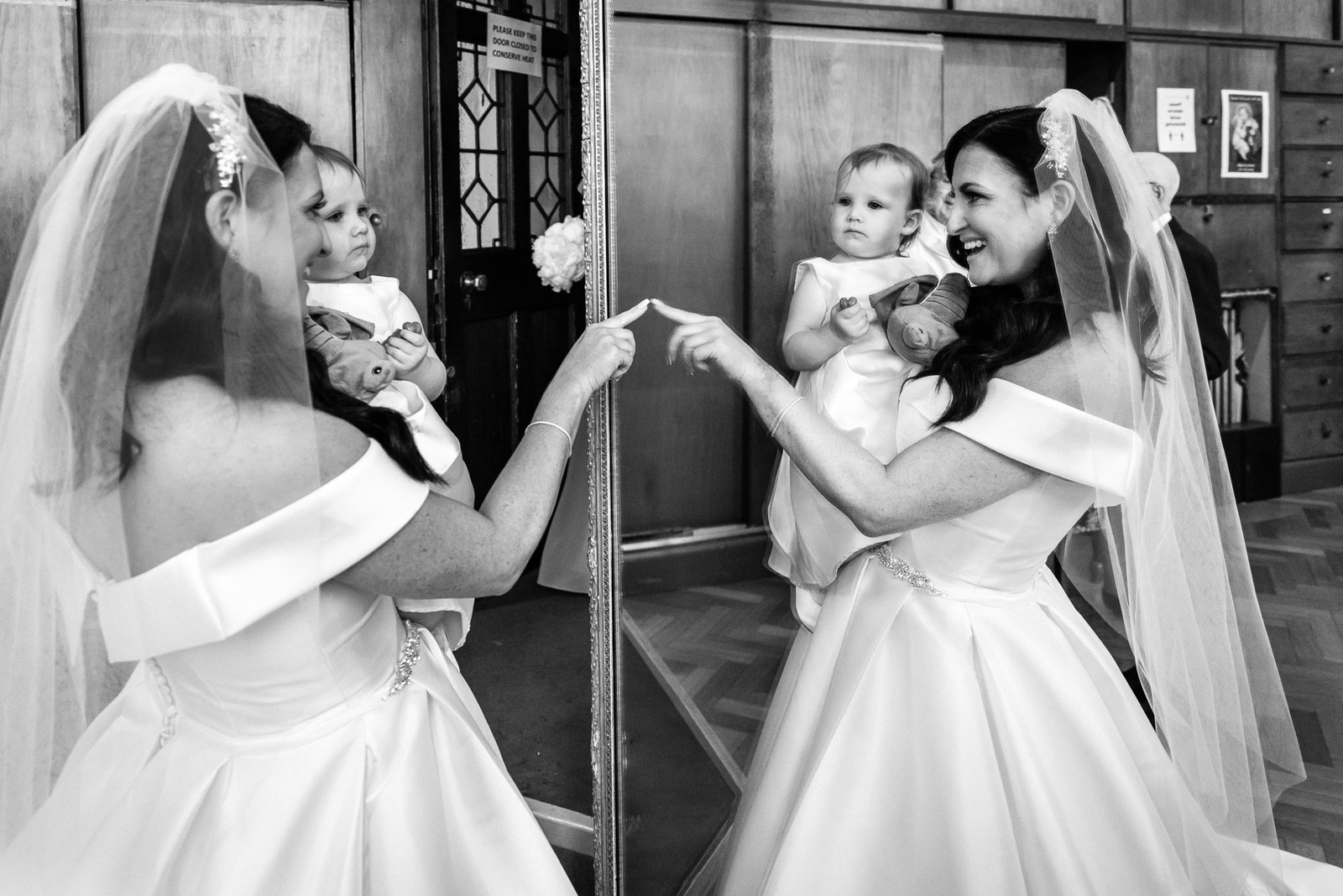 Black-and-white moment of the bride holding a small child beside a tall mirror