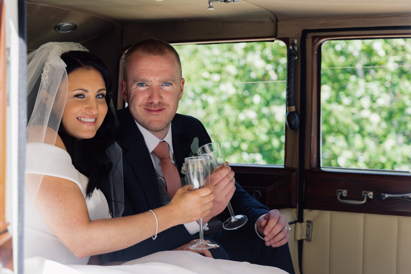 The couple toasting with glasses inside the vintage wedding car