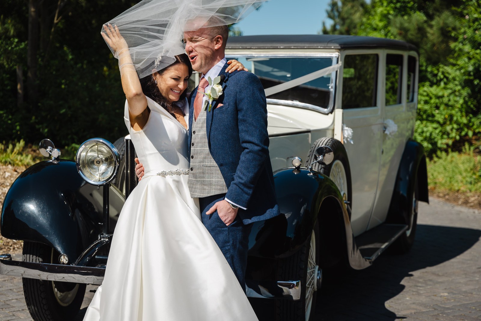 Bride and groom laughing beneath the veil by the vintage car