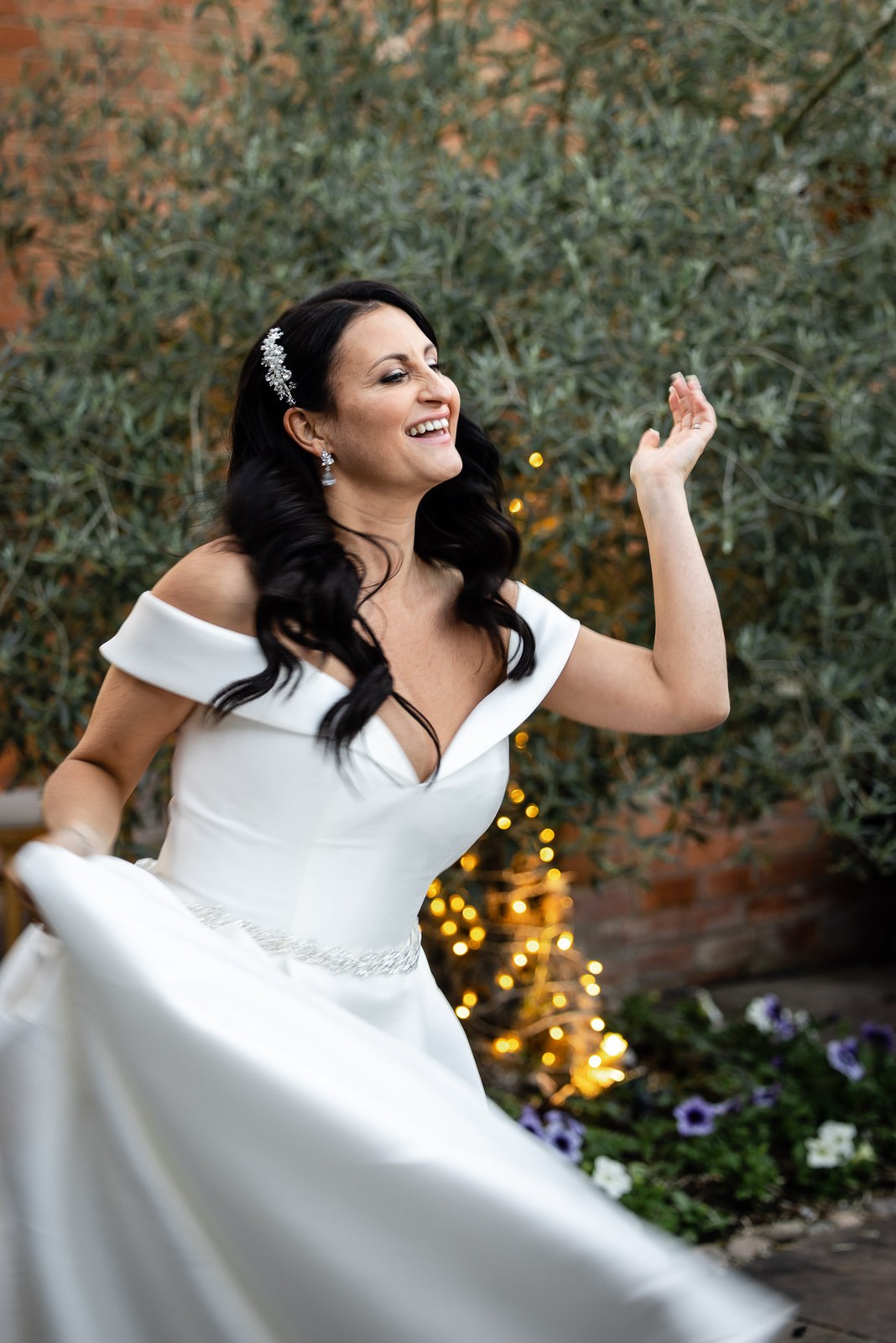 Bride laughing and twirling her gown by a fairy-lit flowerbed at dusk