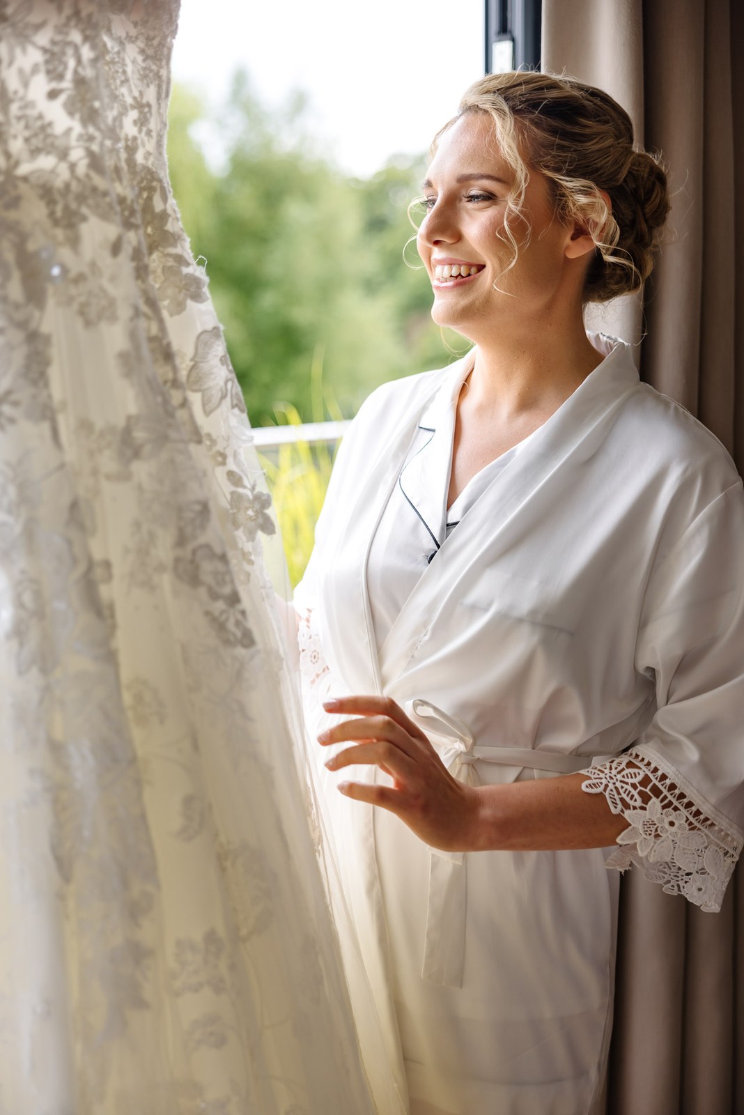 Bride in a robe admiring her lace gown hanging by the window