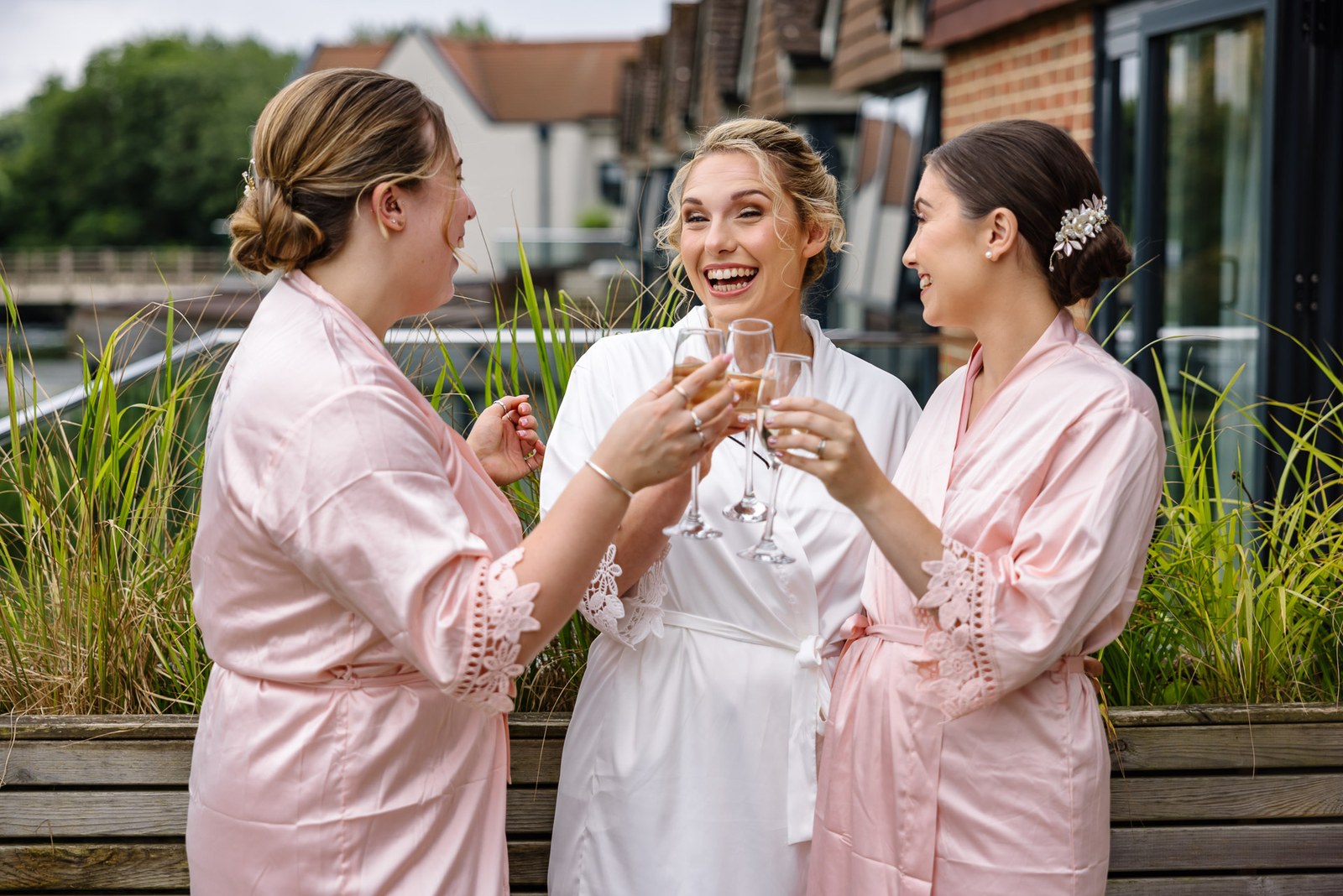 Bride in white and bridesmaids in pink robes toasting on the riverside terrace at The Swan at Streatley
