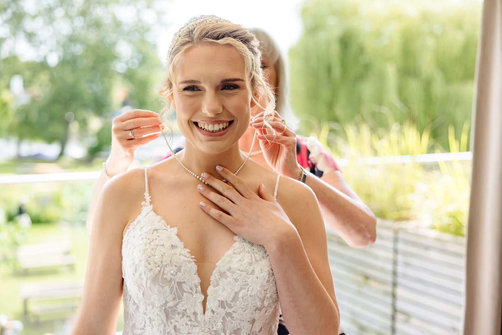 Bride laughing as a helper fastens her necklace outdoors
