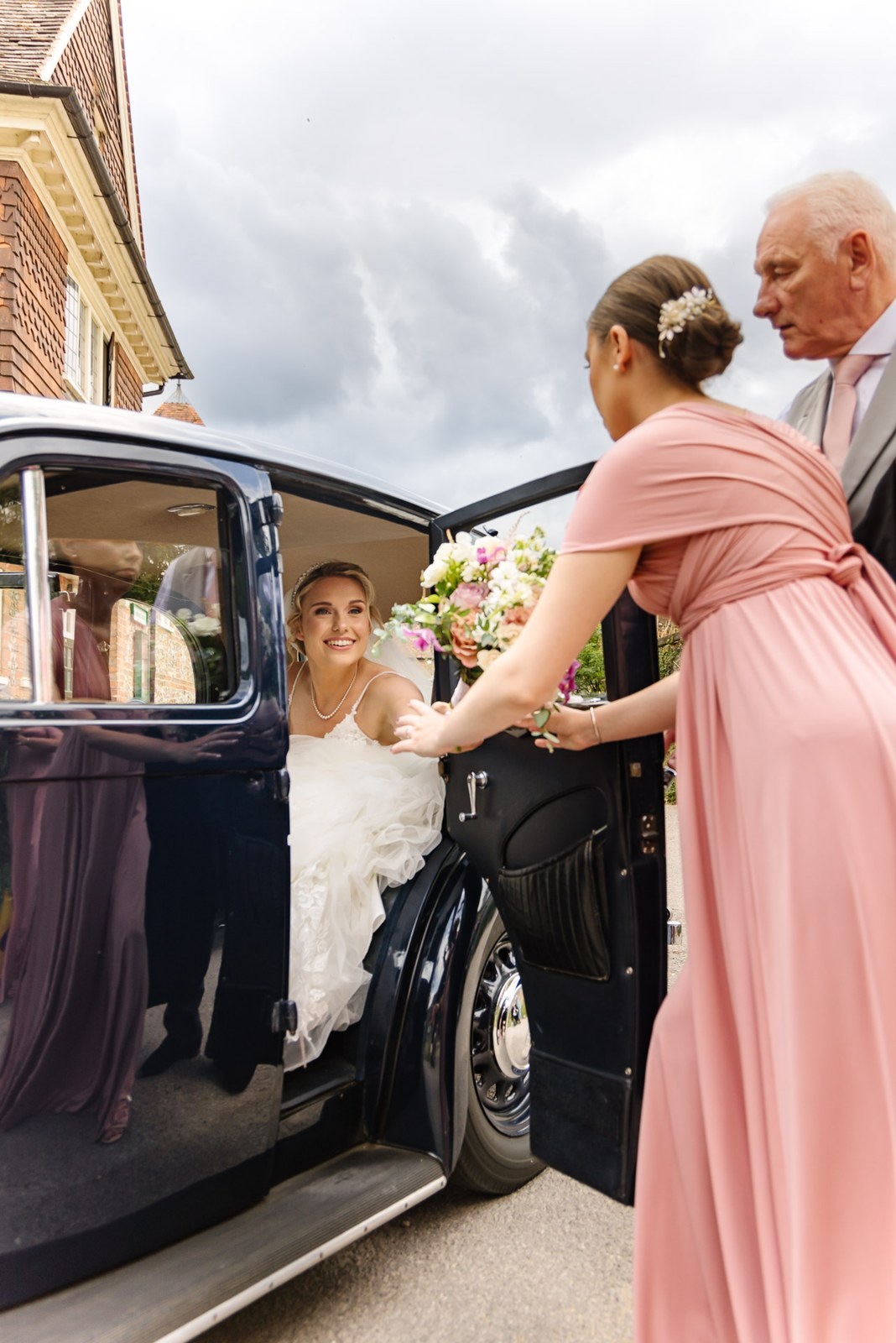 Bride stepping from the wedding car, helped by a bridesmaid and her father