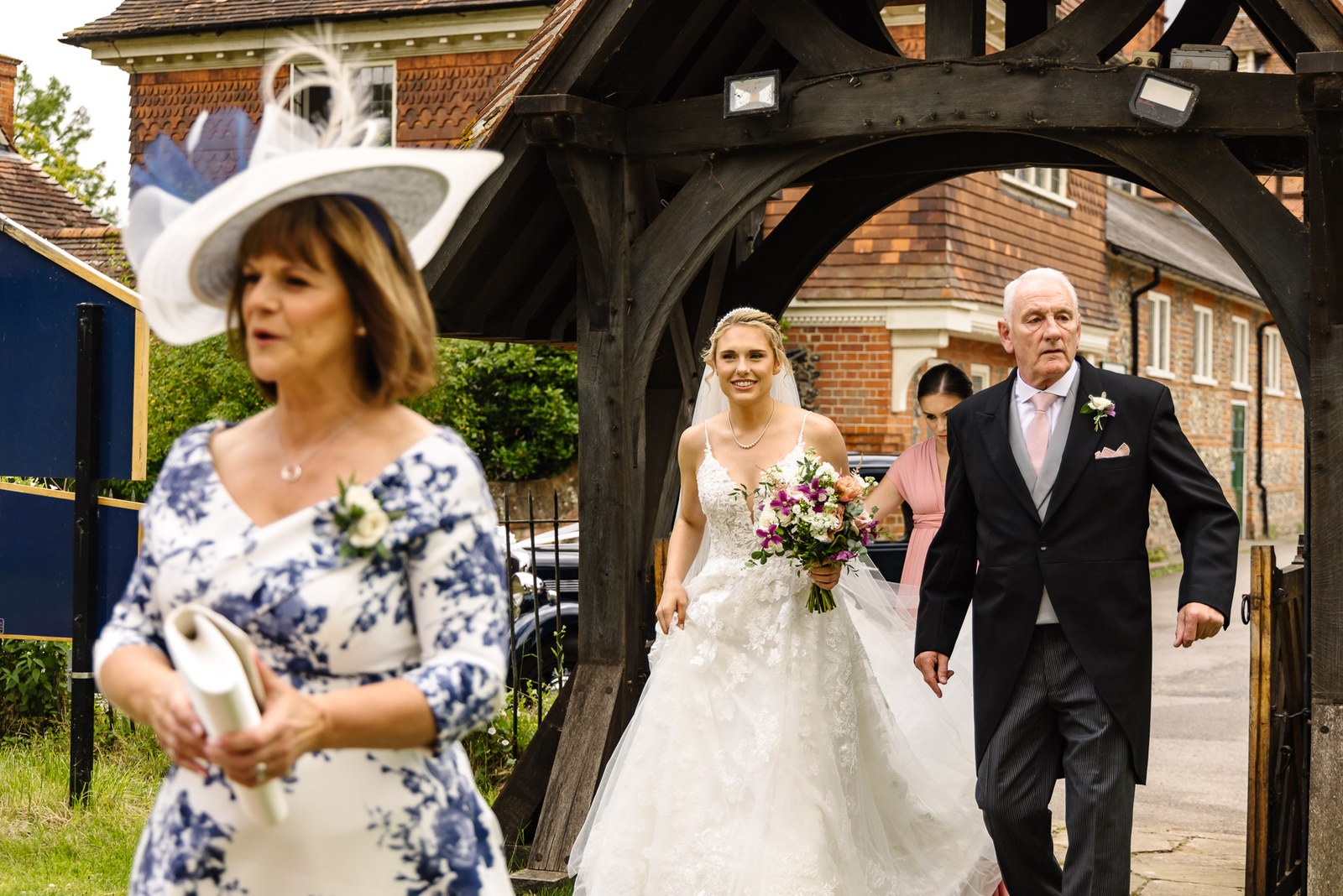 Bride walking through a lychgate on her father's arm, her mother leading