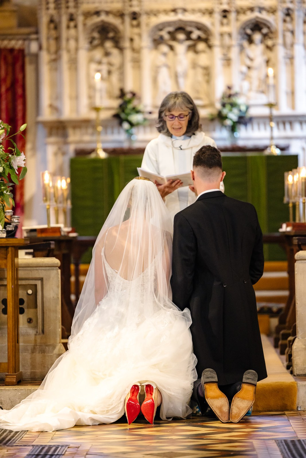 Bride and groom kneeling at the altar before the priest, red-soled shoes showing