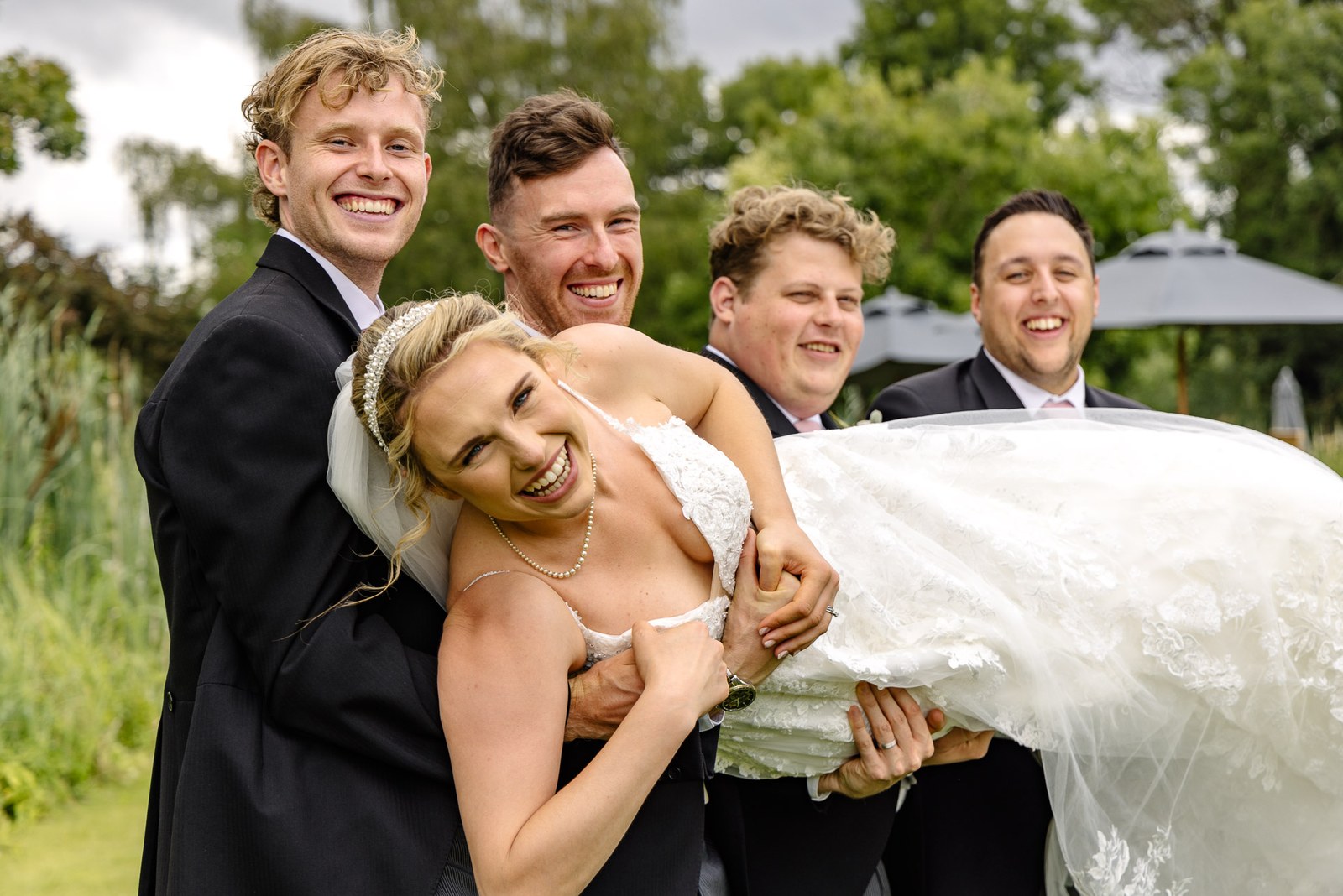 The groom and his groomsmen playfully carrying the laughing bride across the lawn