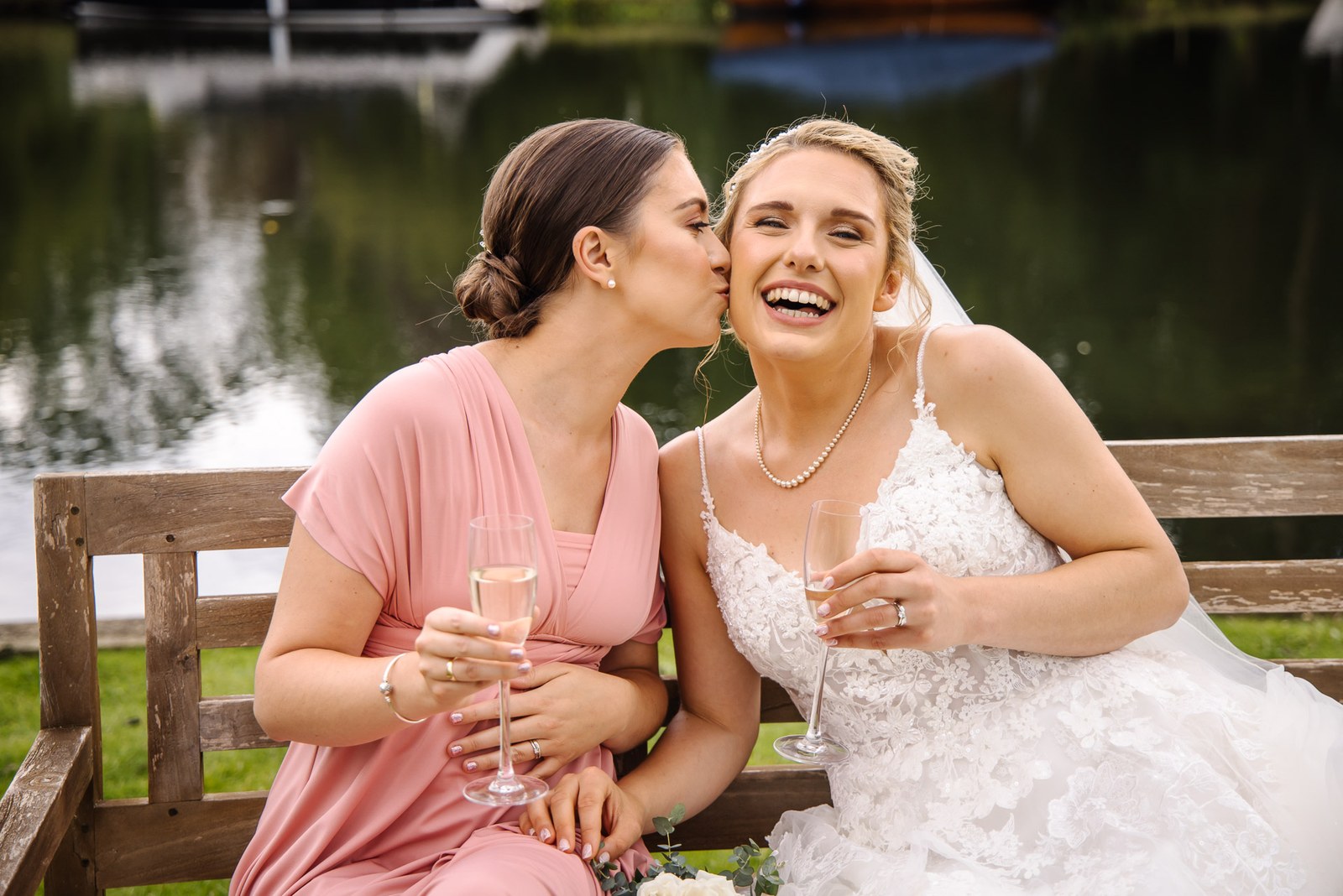 A bridesmaid kissing the bride's cheek on a riverside bench at The Swan at Streatley