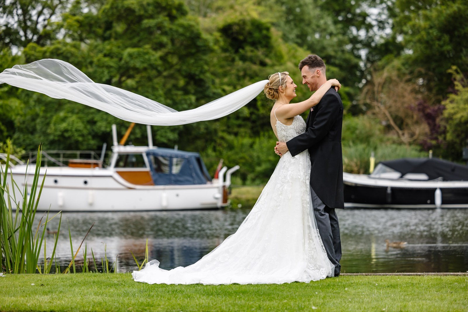 The couple embracing beside the Thames at The Swan at Streatley, veil billowing