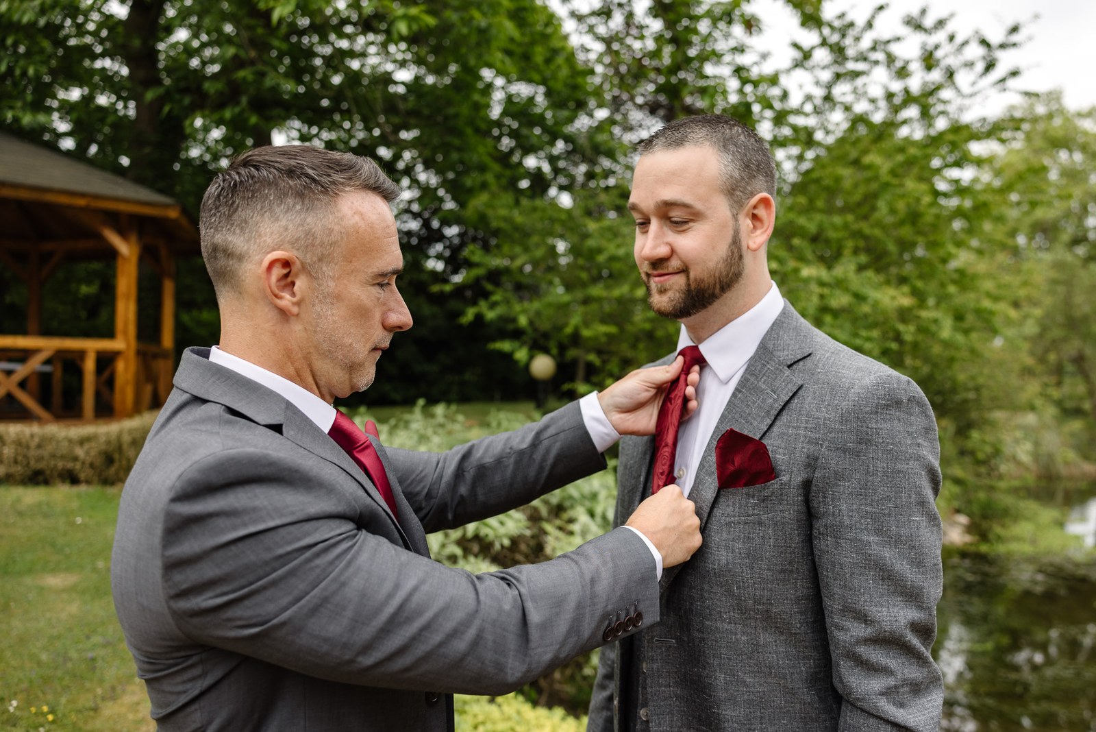A groomsman adjusting the groom's red tie in the gardens