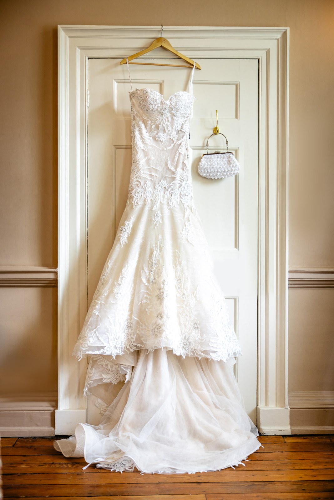 The lace wedding gown hanging on a panelled door with a beaded bag